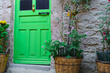 A Greek Style House With a Green Door With Stone Walls And a Fresh Tree And Colorful Flowers in Wicker Pots.