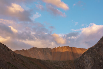 Fototapeta premium Dramatic landscape with sunlit wide sharp mountain ridge under clouds in sunset colors at changeable weather. Atmospheric mountain scenery with large sharp rocks on ridge top under sunset cloudy sky.