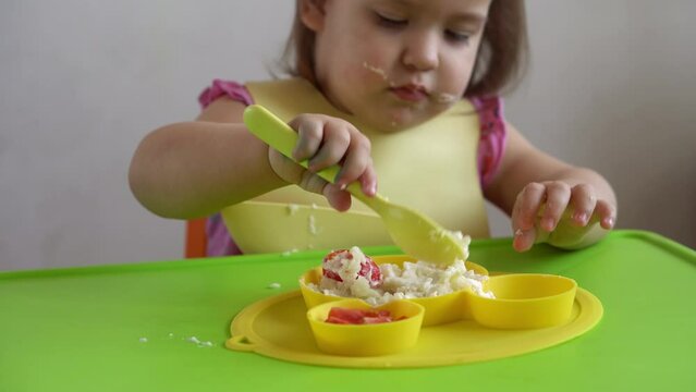 Close up of lovely baby girl eating have breakfast eggs vegetables sausage by herself at home. Adorable possitive happy toddler child eats with dirty face at table indoor. Food, nutrition healthy life