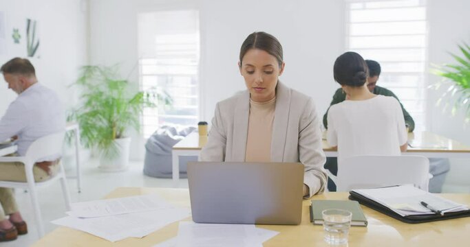Business woman working on laptop with paperwork in a busy office with her colleagues in the background. Focused entrepreneur browsing the internet while planning ideas at her desk in a startup agency