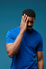 Vertical portrait of handsome black man wearing blue fitting shirt against vibrant blue background and covering face with hand