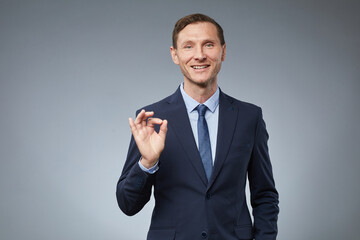 Waist up portrait of smiling Caucasian businessman showing OK sign while standing against grey background in studio, copy space