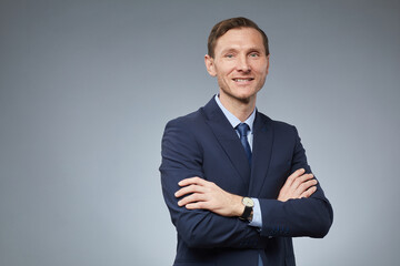 Waist up portrait of smiling Caucasian businessman standing with arms crossed and looking at camera against grey background, copy space
