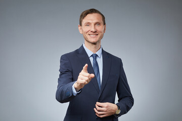 Waist up portrait of smiling Caucasian businessman pointing to camera while standing against grey background, copy space