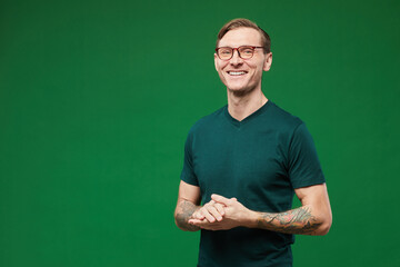 Waist up portrait of tattooed man wearing green shirt and glasses against vibrant green background in studio and smiling at camera, copy space