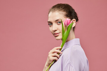 Minimal portrait of elegant young woman holding single pink tulip while standing against vibrant pink background, copy space