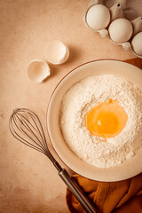 Food background, concept, plate with wheat flour, broken egg, whisk, kitchen towel, on a beige table, top view, rustic,