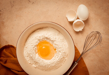 Food background, concept, plate with wheat flour, broken egg, whisk, kitchen towel, on a beige table, top view, rustic,