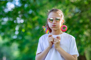 A teenage girl in sunglasses holds lollipops in the form of a pink donut and ice cream. Walking child in the summer park. Horizontal photo of people.