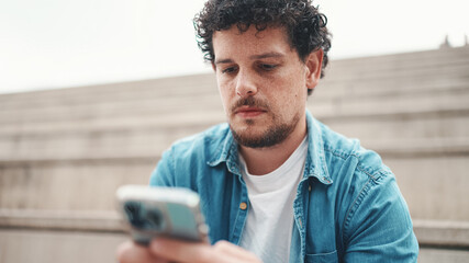  young bearded man in denim shirt sitting on high steps and using mobile phone. Man playing on smartphone