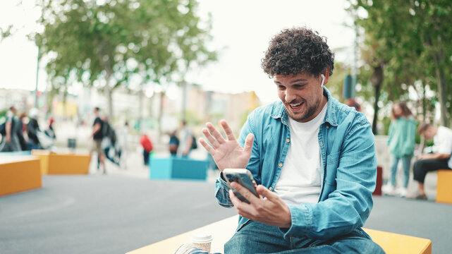 Clouseup, young bearded man in denim shirt sitting in wireless headphones making video call on mobile phone on busy street modern city background.