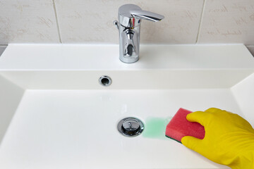 Hand in yellow protective gloves cleans the sink in the bathroom with a sponge and detergent
