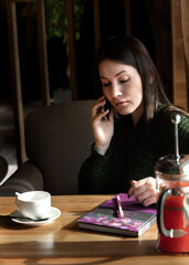 young beautiful woman talking on the phone at a table in a cafe near the window, sea buckthorn tea and a notepad in front of her