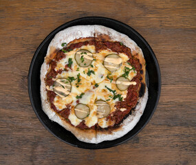 Food. Top view of a pizza with minced meat, tomato sauce, grilled cucumber, garlic aioli and chives, in a black pizza pan on the wooden table. 