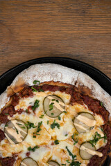 Food. Top view of a pizza with minced meat, tomato sauce, grilled cucumber, garlic aioli and chives, in a black pizza pan on the wooden table. 