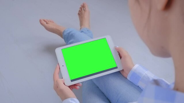 Green screen, mock up, entertainment, copyspace, freelance, template, leisure time, technology concept. Woman sitting on floor and looking at white tablet computer device with blank green display