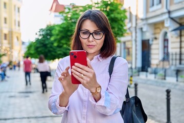 Middle aged smiling woman looking into smartphone on city street