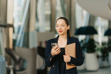 A female realtor is holding a laptop and a smartphone. A pretty woman is standing in the modern lobby. A businesswoman is leaving her office in a contemporary interior.