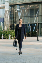 A full length photo of a female realtor in a blazer who is walking with a laptop between skyscrapers.