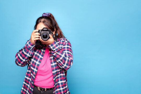 Young curvy Latina woman taking a photo with a reflex camera in a studio, isolated on blue background.