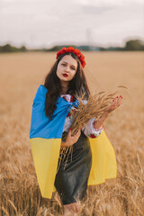 Young sad girl shrouded in flag waiting for victory in the field wearing Ukrainian national embroidered shirt with red wreath and holds wheat bouquet. Concept of Ukraine freedom and stop the war