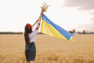 Young girl waiting for victory in the field wearing Ukrainian national embroidered shirt with red wreath and holds up flag and wheat bouquet . Independence Concept, freedom of Ukraine and stop the war