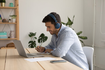 Man sit at desk looks at laptop screen listens to online teacher through wireless headphones,...