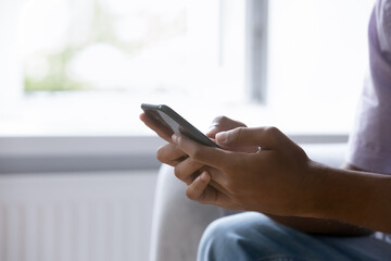 Close up cropped shot African guy hands holding modern smartphone sit on sofa at home. Electronic services buyer, retail e-commerce client, buy goods, search information on internet, e-dating concept