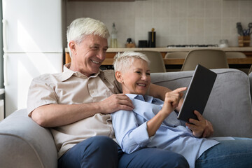 Older couple relax on sofa use digital tablet, choose goods, buy services feel happy enjoy easy comfort delivery e-services, make order, rest at home with modern tech. E-commerce, e-shopping concept