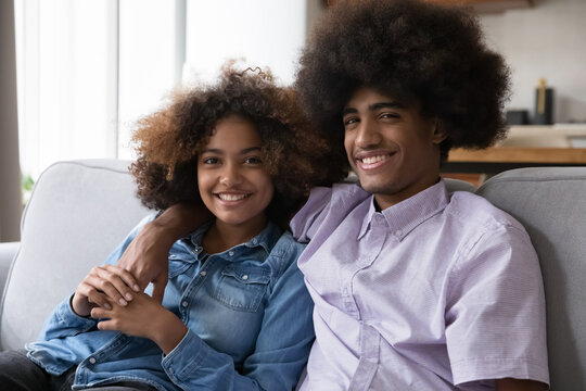Portrait beautiful African girl and guy, romantic teenage couple with natural curly hair smile look at camera pose sit on sofa at home. First love, romance, date, relationships, gen Z teen-age concept