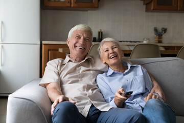 Laughing older couple relax on sofa spend carefree weekend together at home, hold remote control...