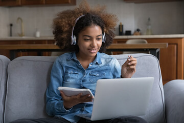 Fototapeta na wymiar Attentive African teen-age girl studying at home, listens audio through wireless headphones, make task use laptop, engaged in easy and effective distancing on-line class. Video call, e-learn concept