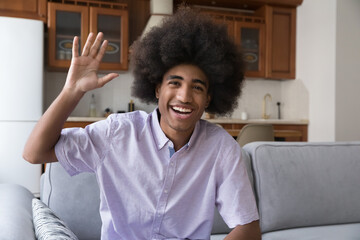 Happy teenage African curly-haired guy sit on couch smile look at camera, wave hand greets friend start videocall. Electronic dating, distance communication, young gen and modern tech usage concept