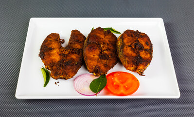 Fish fry arranged beautifully and garnished with onion, tomato slices and green leaves on a white ceramic plate with a grey textured background.