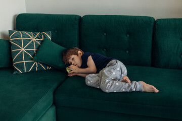 Little boy in sleepwear lying on big green modern sofa in living room and sucking finger. Bad...