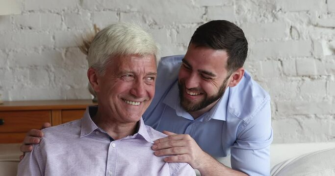 Happy cheerful grown son and old grey haired dad looking at camera with toothy smile, laughing. Positive man touching, holding mature fathers shoulders with care, love, support. Head shot portrait