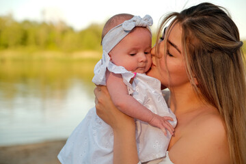 a beautiful girl in a white dress with a newborn baby is sitting on the beach, on the sand.