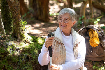 Attractive senior woman sitting in the forest holding a walking cane, elderly lady traveling in nature with backpack enjoying healthy lifestyle and freedom