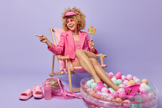 Happy positive curly haired woman points into distance holds sweet lollipop wears sunglasses and pink formal jacket sunbathes at beach poses on deck chair surrounded by inflated pool slippers net bag