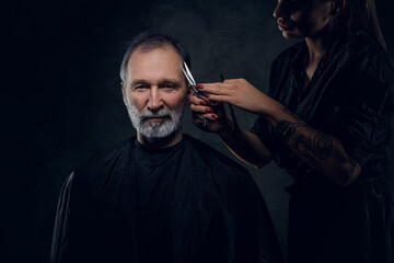 Portrait of woman hairdresser cutting hairs of her elderly customer against dark background.