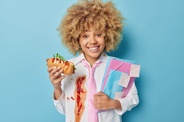Happy curly woman office worker has lunch during work break eats tasty hot dog with ketchup dressed...