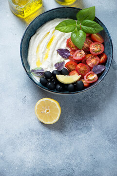 Bowl of greek tarama dip with olives, cherry tomatoes and fresh basil leaves, elevated view on a light-blue stone background, vertical shot with space