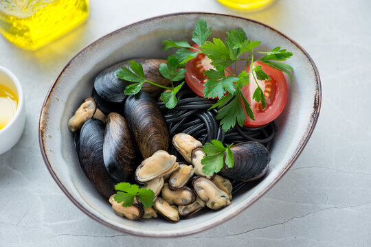 Bowl of mussels with black spaghetti, tomatoes and parsley, studio shot on a light-grey stone background, middle close-up