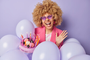 Overjoyed woman with blonde curly hair exclaims loudly holds delicious festive cake going to blow out candles make wish celebrates anniversary on party wears fashionable clothes stands near balloons