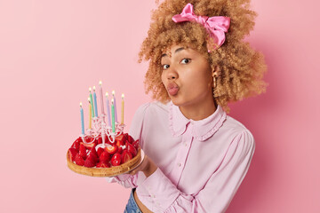 Pretty young woman with curly hair keeps lips folded has romantic mood holds strawberry cake celebrates birthday dressed in fashionable blouse and bowtie on head isolated over pink background