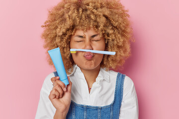 Positive curly haired woman holds tube of toothpaste and toothbrush on folded lips foolishes around...