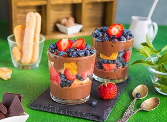Summer dessert in layers in a glass with fresh strawberries, honeysuckle, biscuits and chocolate custard on a green concrete background. Desserts without baking