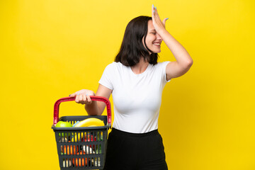 Young woman holding a shopping basket full of food isolated on yellow background has realized something and intending the solution