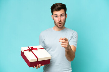 Young caucasian man holding a gift isolated on blue background surprised and pointing front