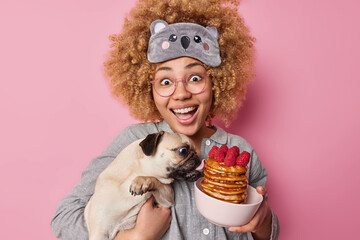 Positive woman with curly hair dressed in nightwear carries pedigree dog which licks delicious...